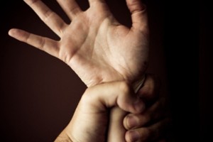 A male hand grips a female hand around the wrist, her fingers are spread, the picture suggest dominance. It is on a black backgound